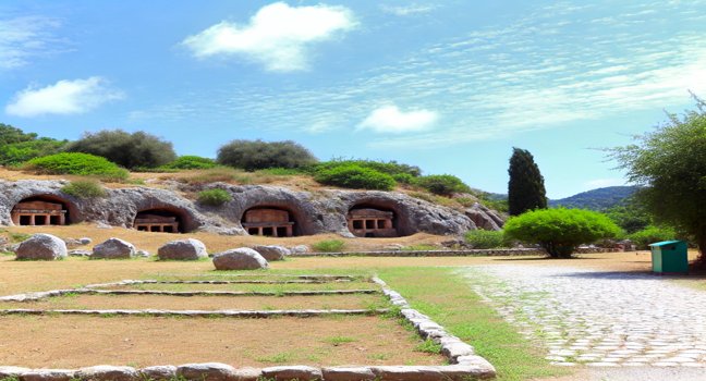 A scenic view of Fethiye's historical places, featuring ancient rock-cut tombs carved into cliffs, surrounded by lush greenery and pathways under a bright blue sky