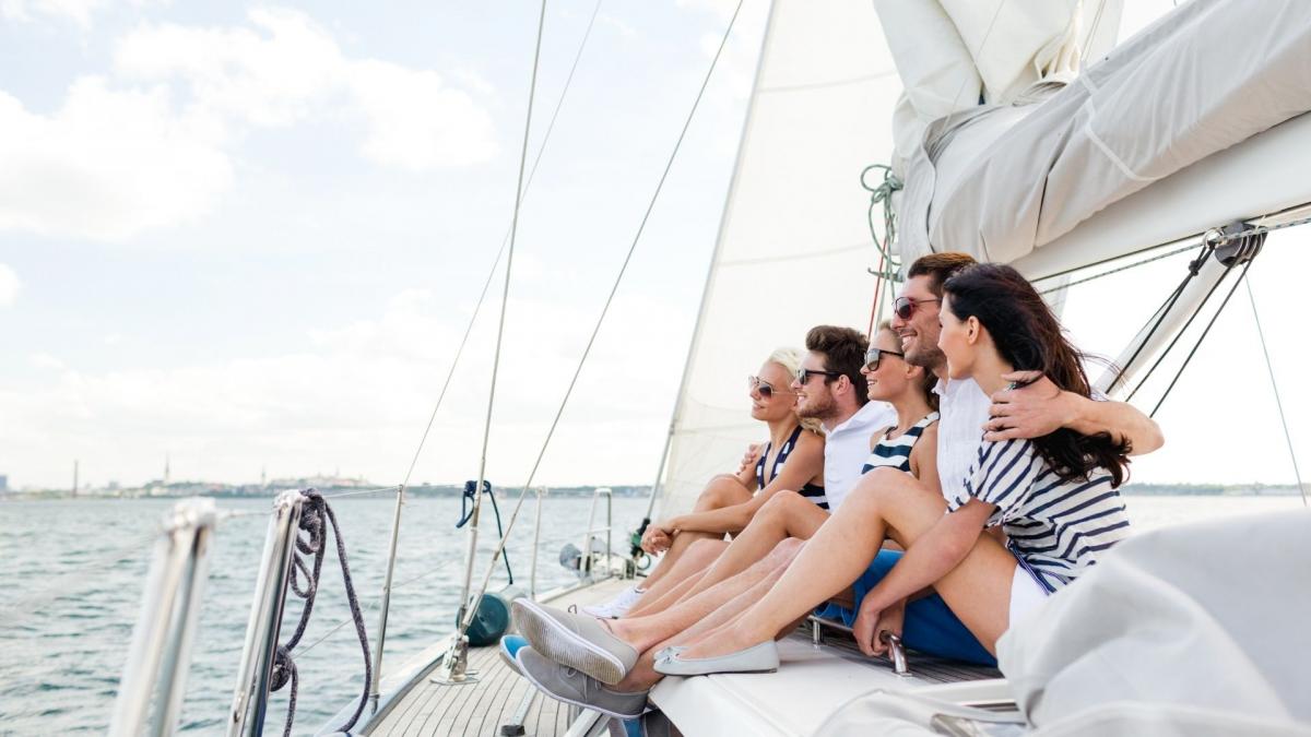 Eine Freundesgruppe genießt ihren Sommerurlaub auf einer Segelyacht, lächelnd und entspannt mit Blick auf das weite Meer und die Küste im Hintergrund