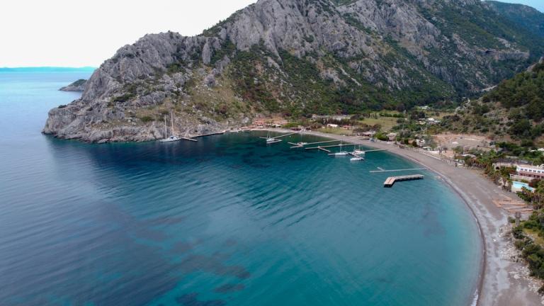 A serene view of Çiftlik Bay with calm turquoise waters, rugged cliffs, and a few anchored boats by the shoreline, framed by a mountainous landscape