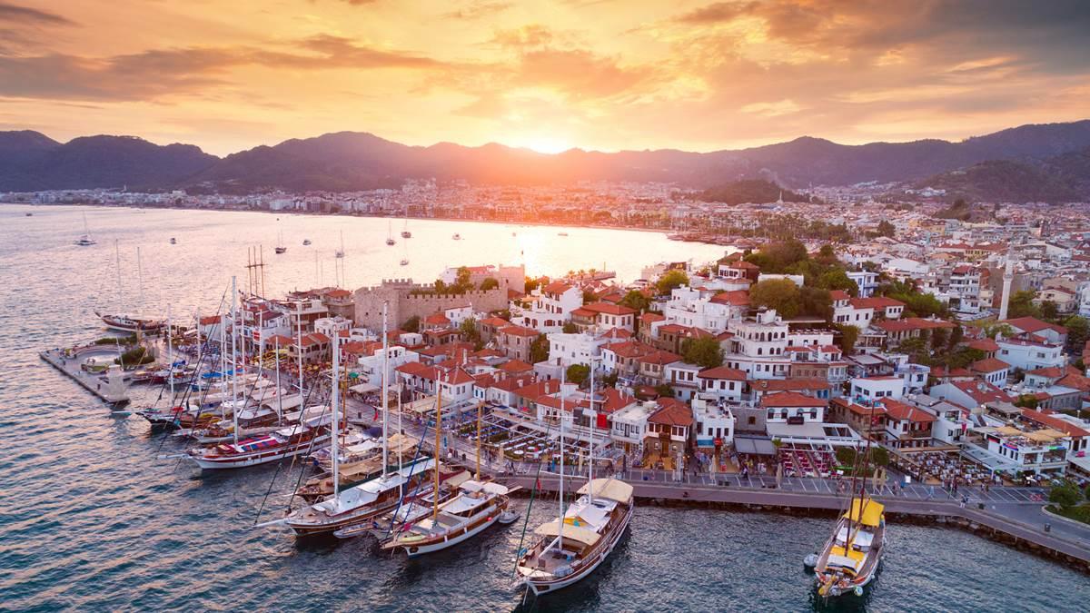 A beautiful view of Marmaris showcasing its bustling harbor with boats and yachts, white and terracotta-roofed buildings, and rolling hills illuminated by the warm glow of a sunset over the calm sea