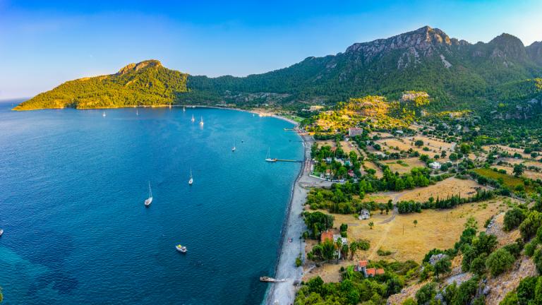 An aerial view of Kumlubük Bay, showing deep blue waters, anchored boats, a stretch of beach, green hillsides, and scattered buildings near the shoreline, with mountainous terrain in the background