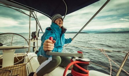 Eine Frau in warmer Kleidung steuert eine Yacht auf offener See. Sie trägt eine Jacke und eine Mütze, während sie das Ruder festhält und den Blick auf das winterliche Meer richtet