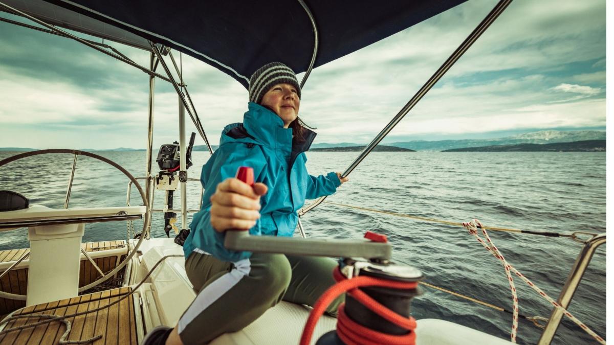 Eine Frau in warmer Kleidung steuert eine Yacht auf offener See. Sie trägt eine Jacke und eine Mütze, während sie das Ruder festhält und den Blick auf das winterliche Meer richtet