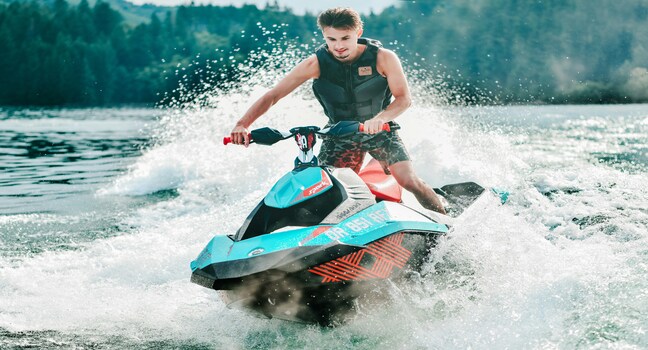 A smiling young man riding a blue jet ski fast across a scenic forest lake