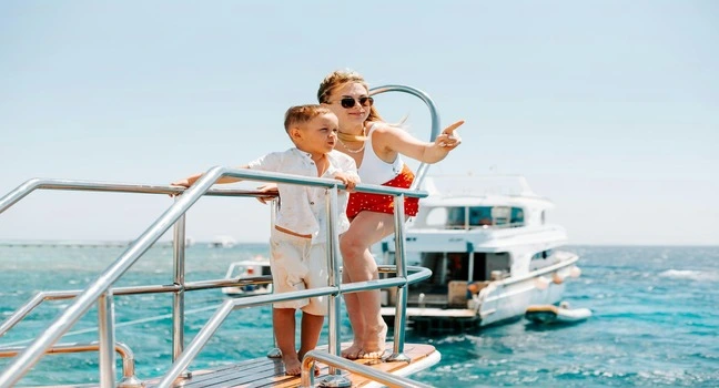 A woman and child on a yacht deck enjoying a sunny day with a luxury boat in the background