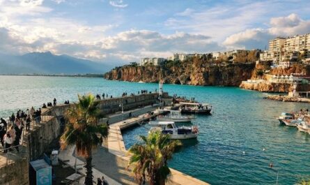 Hafenbereich mit türkisblauem Meer, Felsklippen, Booten und Spaziergängern an der türkischen Küste