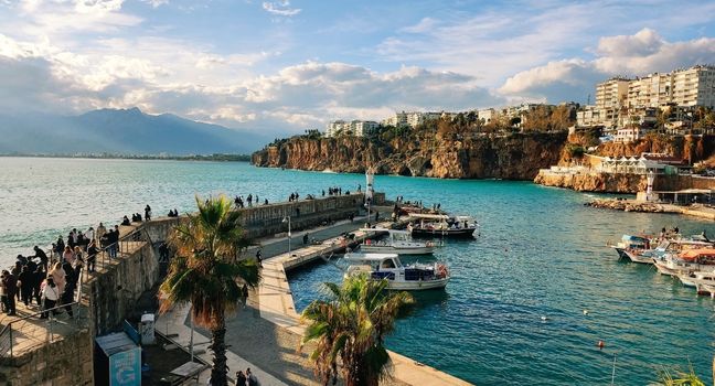 Hafenbereich mit türkisblauem Meer, Felsklippen, Booten und Spaziergängern an der türkischen Küste