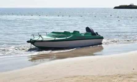 A small green motorboat resting on the sandy shore with calm sea in the background