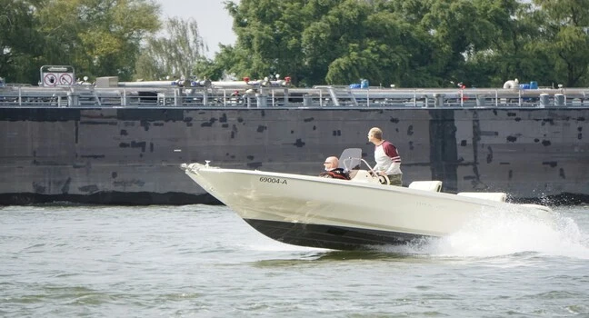 Two men riding a small white motorboat near a large ship on a calm waterway