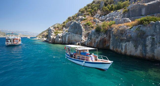 A small tour boat glides through crystal-clear blue waters near rocky coastal cliffs, offering a tranquil and scenic experience