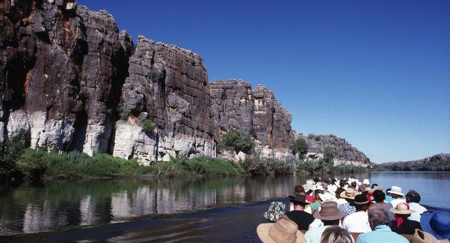 A group of travelers enjoys a serene river cruise surrounded by towering cliffs and lush greenery under a clear blue sky