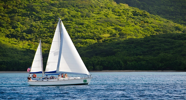A white sailing yacht with raised sails passing a lush green island coast