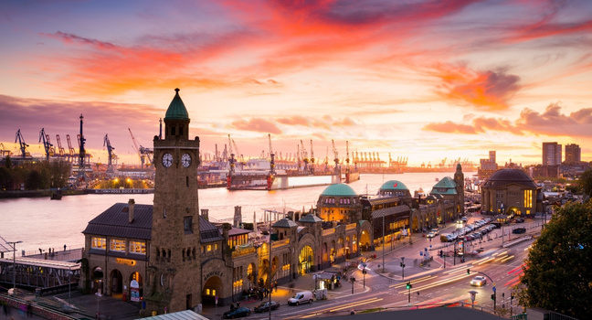 Blick auf die Hamburger Landungsbrücken bei Sonnenuntergang mit Hafenkranen im Hintergrund, aufgenommen entlang der Route einer Hafenrundfahrt
