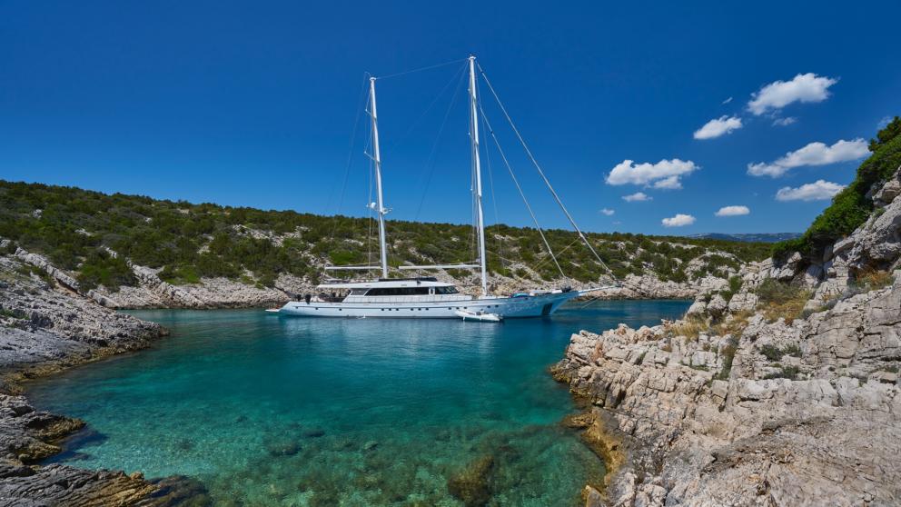 The luxury yacht sits on crystal clear water in a secluded bay, surrounded by rocks under the blue sky.