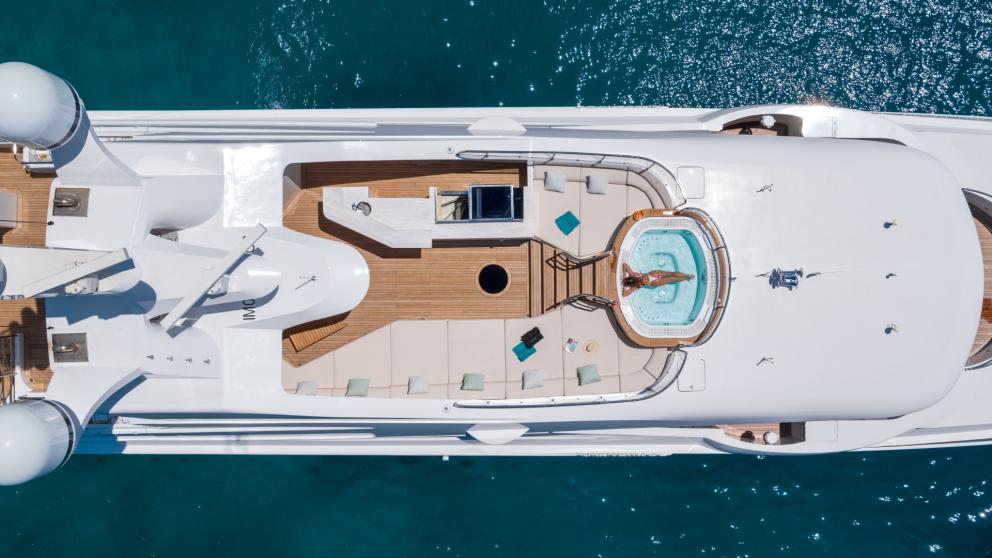 Bird's eye view of a luxury yacht with a whirlpool on deck, surrounded by sun loungers and clear blue water.