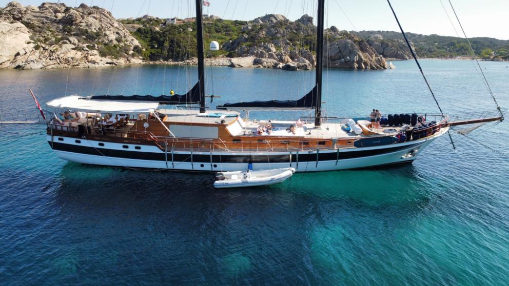 The gulet Elianora anchors in crystal-clear waters off the picturesque, rocky coast of Italy, with a dinghy.