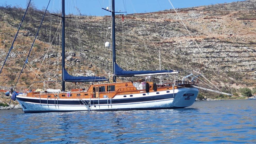The gulet Elianora is anchored in calm waters off a rocky coast in Italy, with an elegant wooden design and black sails.