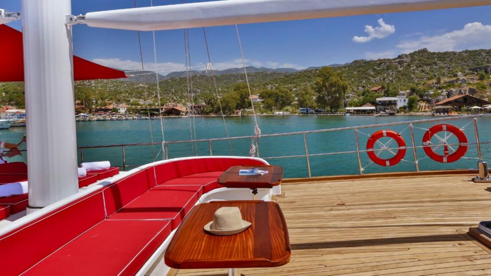 The deck of the gulet Victoria with red upholstered furniture and a view of the turquoise water and the coast.
