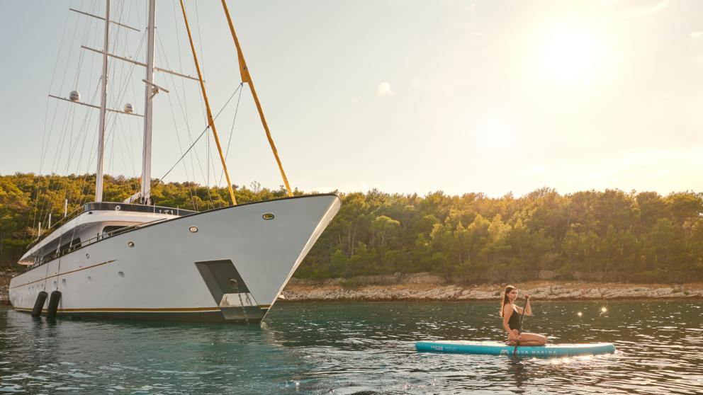 The motor sailor Anima Maris anchored near the coast, with a woman paddleboarding in the water.