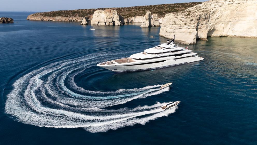 The large luxury yacht is anchored off the rocky coast. Two small speedboats are around it.