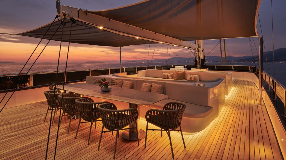 Illuminated deck of Anima Maris at sunset, featuring a dining table and lounge area.