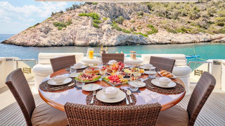 Beautifully set table on the aft deck of a yacht with views of rocky coastline and turquoise water