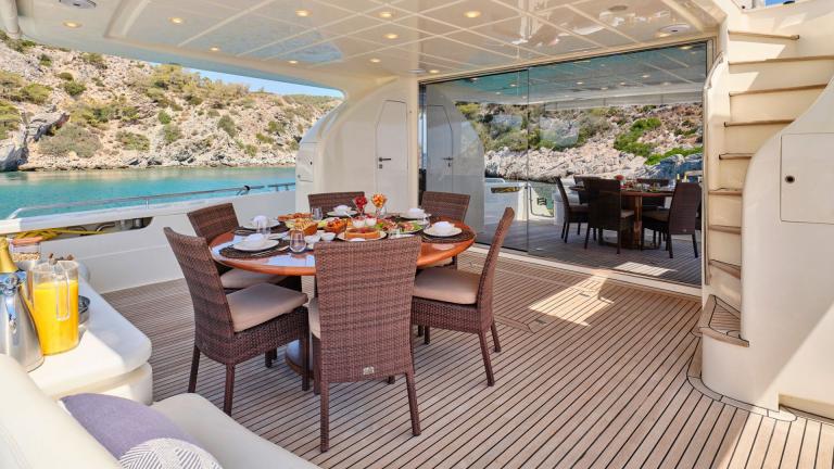 Elegant dining area on the aft deck of a motor yacht with views of rocky coastline and turquoise water