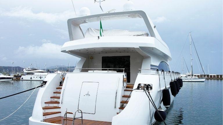  Das Heck einer weißen Luxusyacht, die im Hafen vertäut ist. Die Treppe führt zum oberen Deck, und Fender schützen die Yacht an den Seiten. Im Hintergrund sind weitere Boote und ein bewölkter Himmel zu sehen