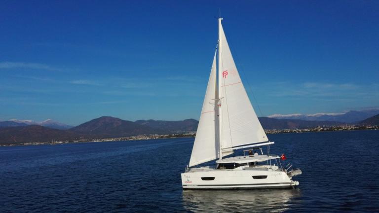 The Hola catamaran sails on calm seas with a backdrop of blue sky and mountains.