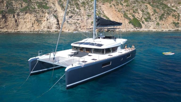 Lagoon 52 F catamaran anchored in clear waters, with a rocky coastline in the background.