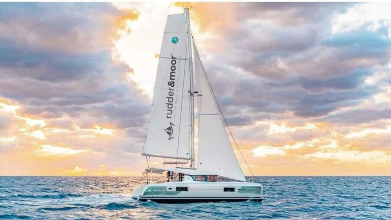 The catamaran Meraki is sailing with its sails open with a view of the clouds.