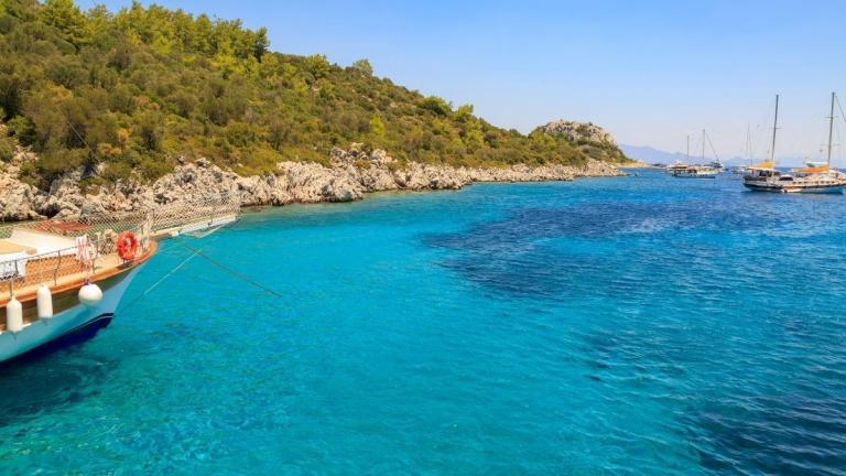 A vibrant scene of crystal-clear turquoise waters along a rocky coastline in Marmaris, Turkey. Several boats and yachts are anchored nearby, creating an inviting atmosphere for a holiday in Marmaris. The lush greenery and calm sea make it a perfect spot for relaxation and exploration