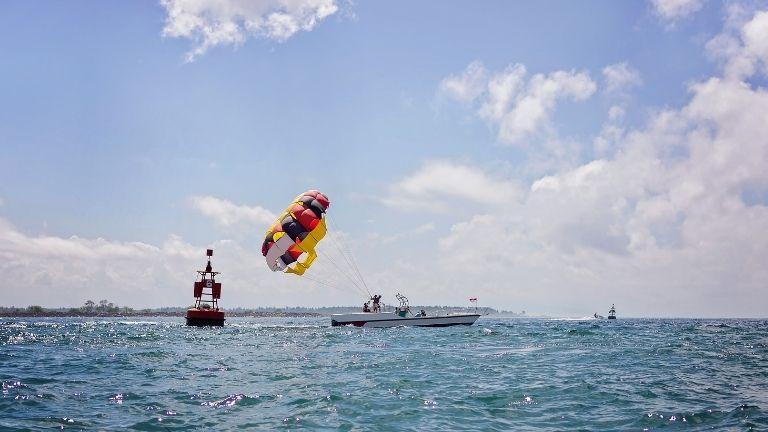 Ein buntes Parasailing-Schirm zieht einen Abenteurer hinter einem Motorboot über das offene Meer, neben einer roten Boje unter blauem Himmel