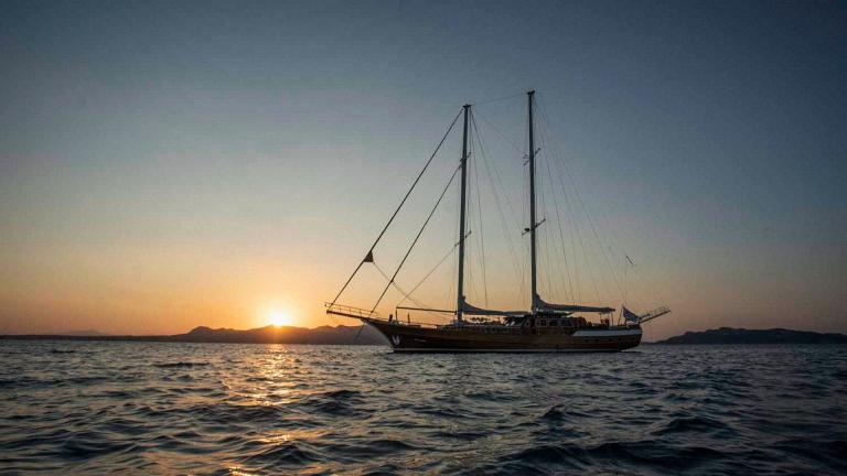 Die Gulet Entre Cielos segelt bei Sonnenuntergang vor der Küste von Athen, silhuettiert gegen den farbenfrohen Himmel.