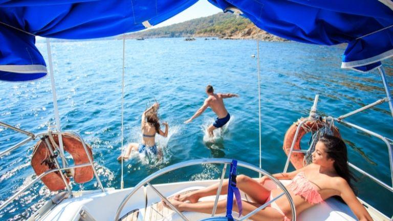 A woman in a bikini is sunbathing on the yacht and the others are jumping into the sea.