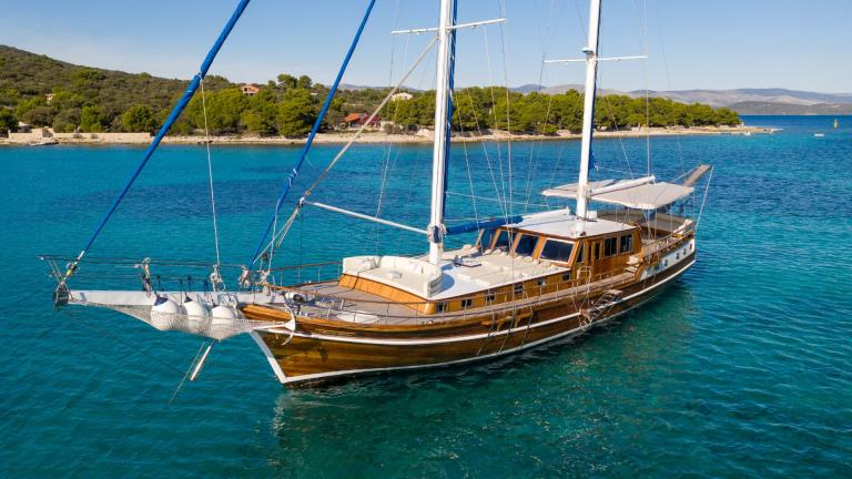 The gulet Andi Star, an elegant wooden sailing yacht, is anchored in the clear, turquoise-coloured water off a wooded co