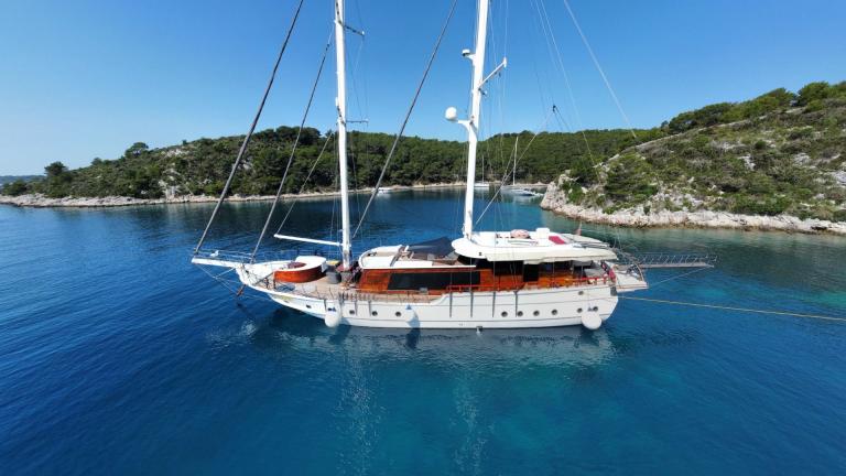A luxurious gulet with six cabins, named Vivere, lies calmly in the clear blue waters off a green coastal landscape in D