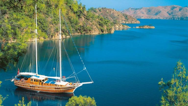 A wooden sailboat is anchored in a tranquil bay surrounded by lush greenery and mountains on the Aegean coast of Turkey.