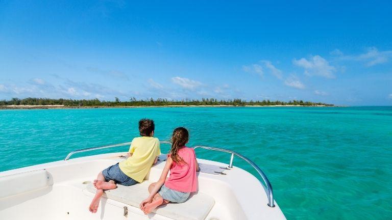 Zwei Kinder sitzen auf dem Bug eines Bootes und blicken auf das türkisfarbene Wasser und die Küstenlinie in der Ferne unter einem strahlend blauen Himmel