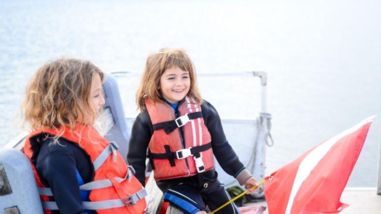 Zwei Kinder tragen Rettungswesten und sitzen fröhlich auf einem Boot. Eines der Kinder hält eine rote Fahne, während sie gemeinsam die Bootsfahrt genießen