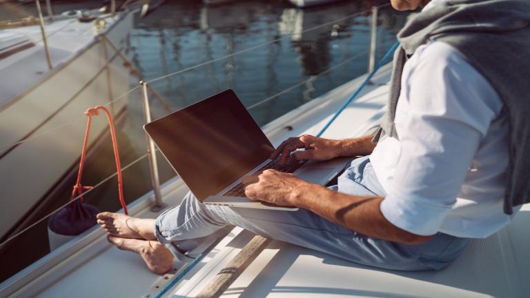 Ein Mann sitzt barfuß an einem Bootssteg und arbeitet entspannt an seinem Laptop.