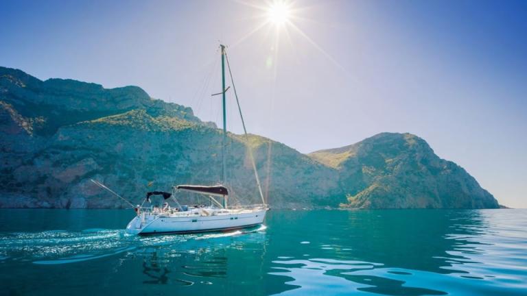 A sailboat glides through the clear blue waters with sunlit mountains in the background on the Aegean coast of Turkey.