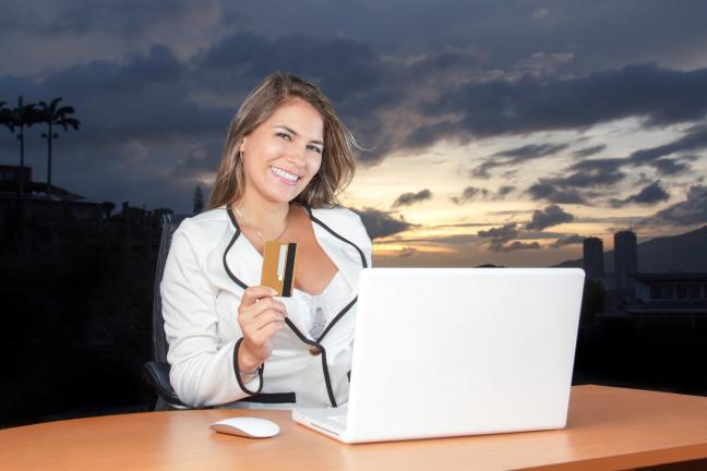 Lächelnde Frau mit Kreditkarte in der Hand sitzt vor einem Laptop im freien. Die Sonne ist bereits untergegangen.