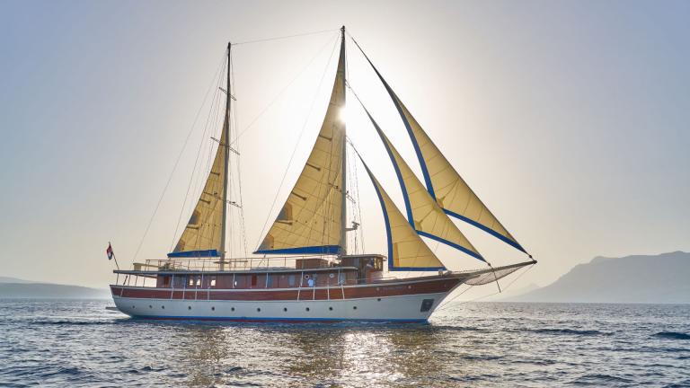 Split'ten 9 kabinli bir gulet, mavi ve sarı yelkenleriyle güneş ışığında yelken açıyor.