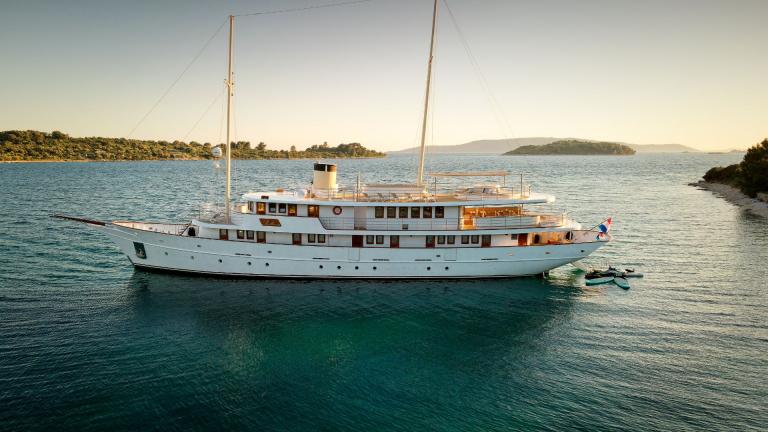 A magnificent white yacht anchored in calm, clear waters, surrounded by scenic islands at sunset.