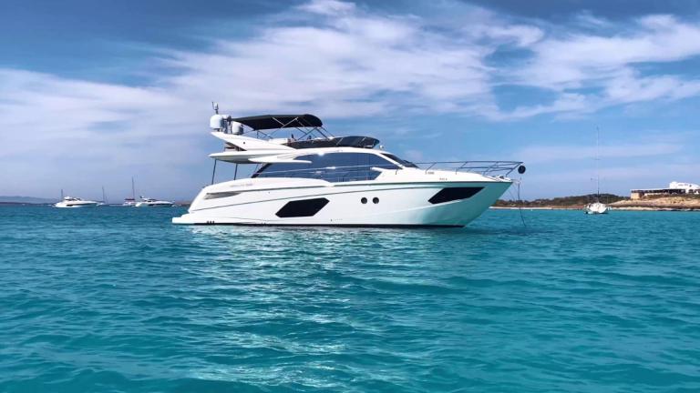 The yacht is stationary with a sleek design and flybridge visible. Calm sea surrounds it near Ibiza.