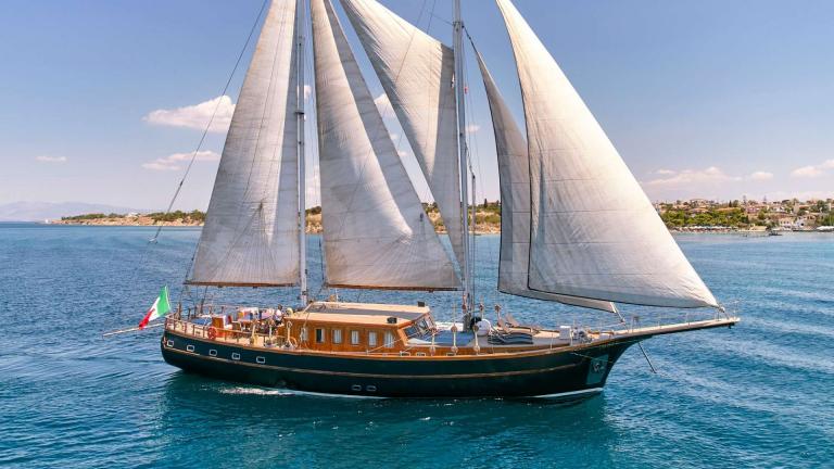 The elegant gulet Myra sails off the coast of Athens, with full sails and a clear sky in the background.