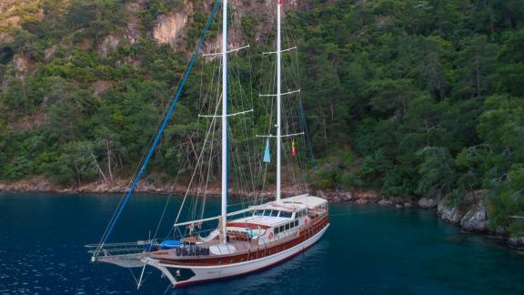 A large turquoise-coloured sailing boat in a bay surrounded by lush forest and rocky slopes, ideal for renting out.