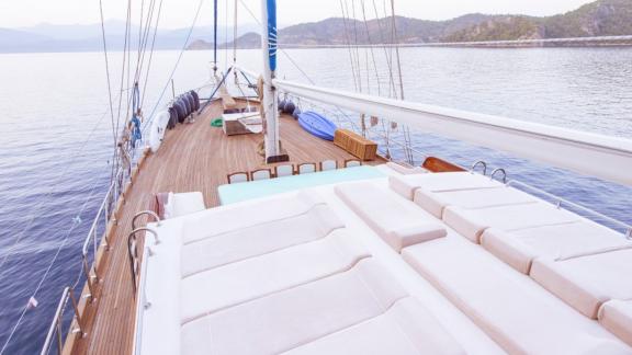 The deck of a sailing boat with sunbathing areas, a dining table and a view of the sea and the coast.