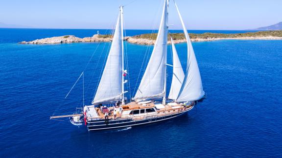 Большая гулета с белыми парусами плывет по спокойной голубой воде у побережья близ Бодрума.